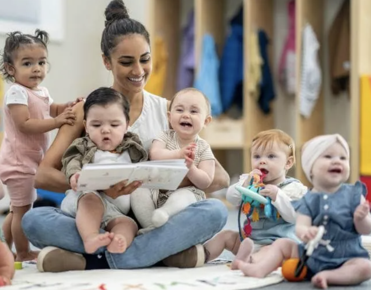 Childcare worker surrounded by babies while sitting on daycare facility floor.