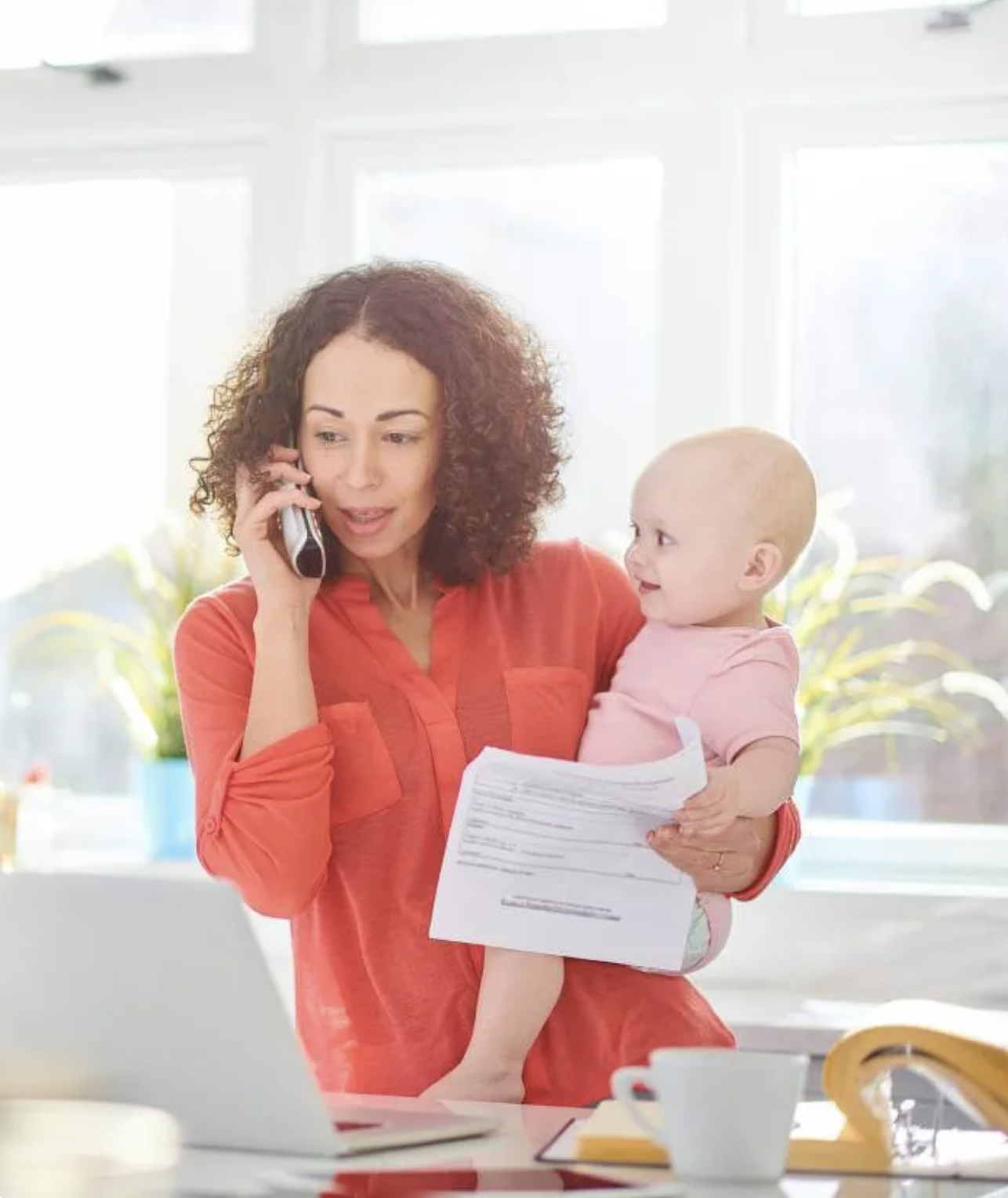 Mother holding a baby while she's looking at a daycare contract and talking on the phone.