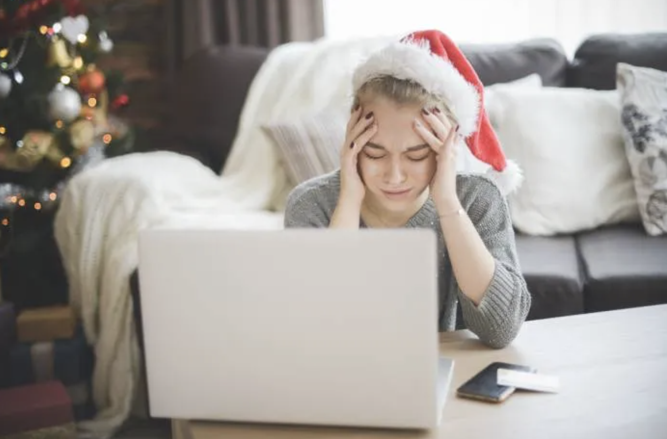 Upset woman wearing a Santa hat sitting on the floor in front of a laptop on a coffee table with Christmas tree in background.