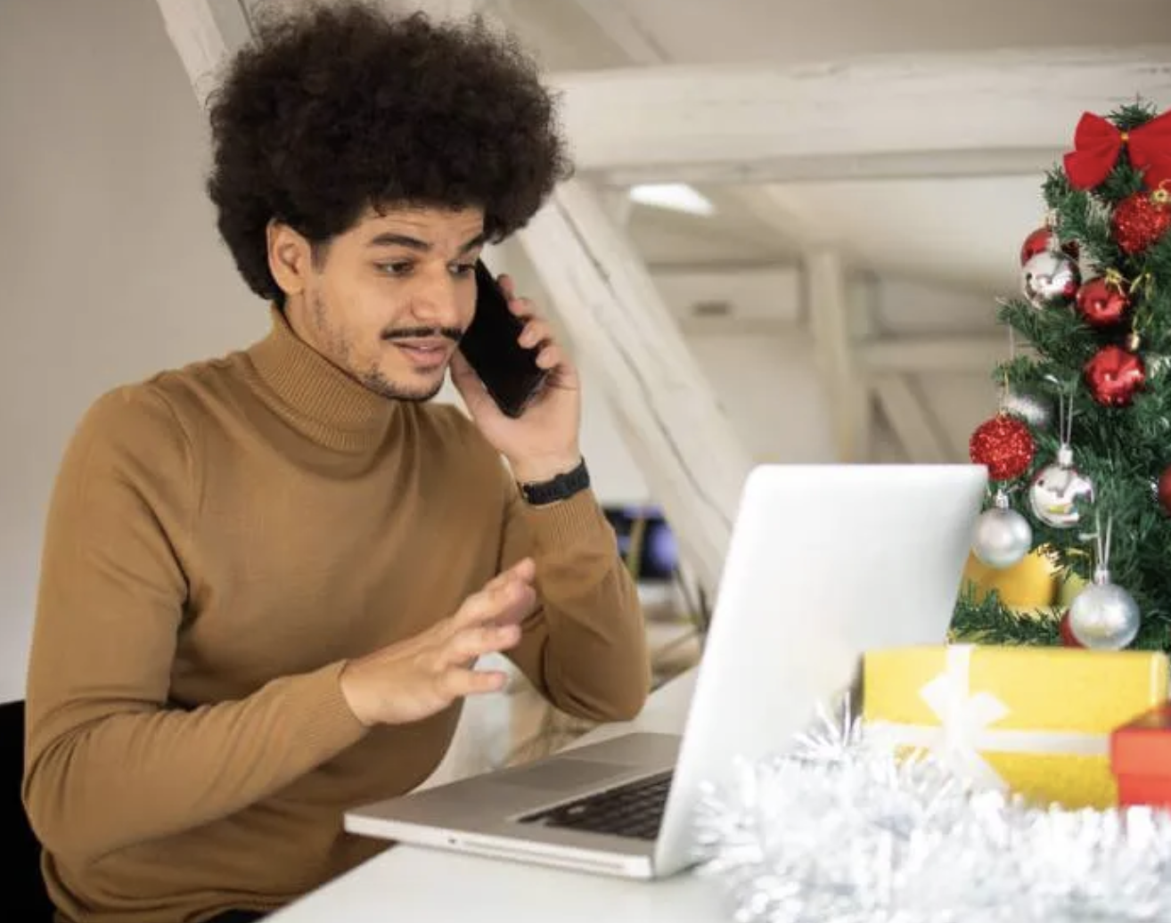 Stressed man talking on phone and looking at his laptop screen hoping he hasn't been scammed. A Christmas tree is in the background.
